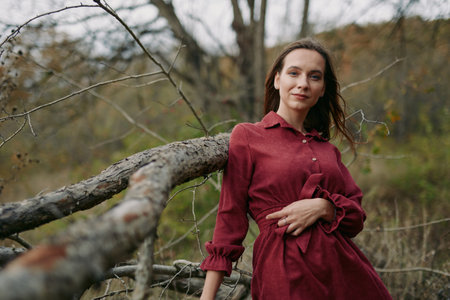 Portrait of a woman in a red dress leaning on a rustic fallen tree, natural expression and relaxed posture conveying authenticity and credibility in a candid outdoor lifestyle scene.の写真素材