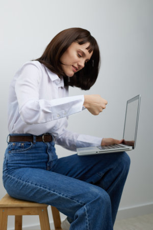 A focused professional woman sits on a wooden stool, using a laptop in a clean indoor setting,...