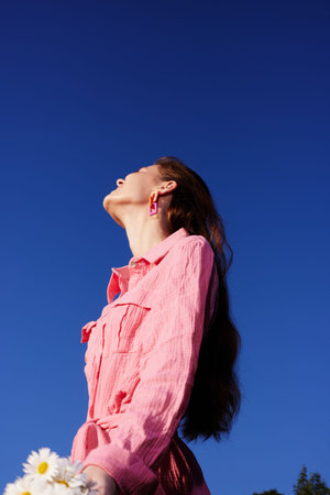 Young woman in pink outfit looking up against clear blue sky, holding daisies, evoking feelings of hope and springtime freshnessの写真素材