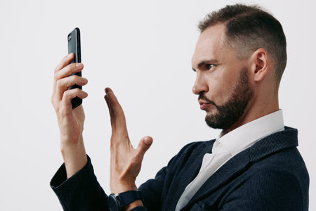 A professional business man stands isolated against a solid light background, with a focused posture, serious expression, and confident gesture as he speaks into a smartphone.の写真素材