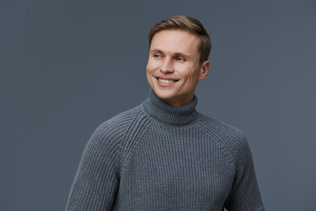 Smiling man wearing gray turtleneck sweater looking away isolated on plain dark gray background in...