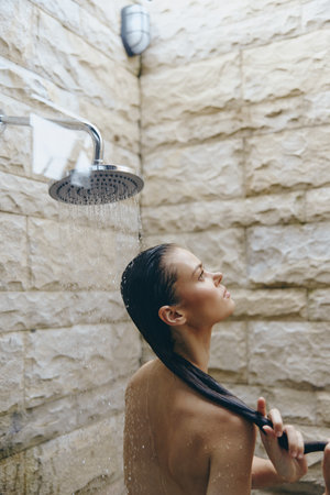 woman showering outdoors with wet hair under rain showerhead on textured stone wall background, natural light and relaxed mood in warm tonesの写真素材