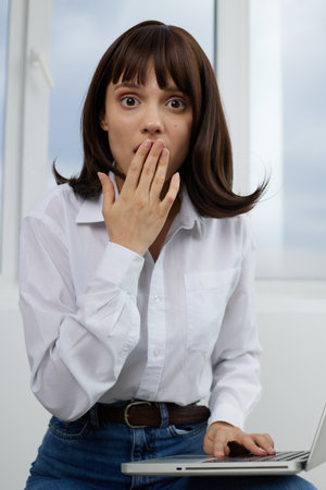 A surprised young woman sits at a desk with a laptop, covering her mouth in disbelief during a...