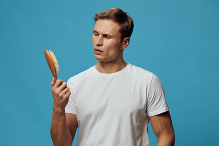 Man looking at hairbrush with puzzled expression, casual white t-shirt, short hairstyle, isolated on blue background, studio shot, people emotion, grooming conceptの写真素材