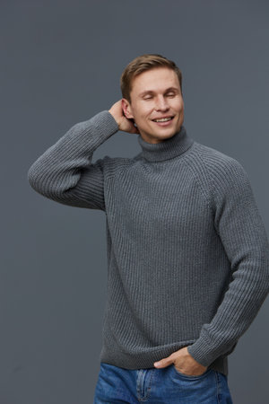smiling man wearing grey knitted turtleneck sweater standing isolated on dark background closed eyes...