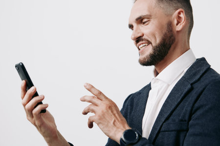 A professional man in a business setting smiles while using a smartphone, isolated on a clean white background. The image conveys confidence, focus, and proactive attitude for corporate communicationの写真素材