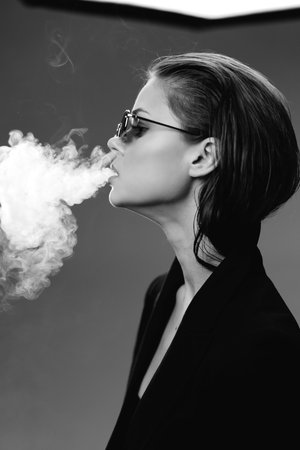 Black and white portrait of a stylish woman exhaling smoke with sunglasses and short hair, creating a moody atmosphere.の写真素材