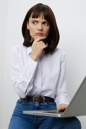 Thoughtful female professional sits with a laptop, chin resting on her hand, studying data and ideas. Casual white shirt and jeans convey a focused, calm workspace.の写真素材