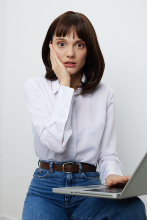 Casual business portrait of a surprised woman seated with a laptop, touching her cheek in a...