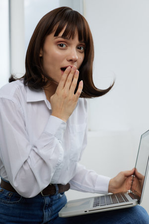 A surprised young woman sits with a laptop on her lap in a bright indoor setting, hand to mouth,...