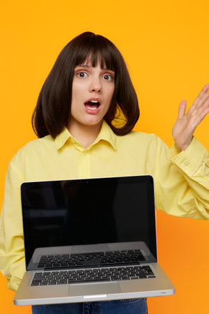 A surprised young woman stands against a bright yellow backdrop, presenting an open laptop with a blank screen, capturing a moment of online discovery and unexpected news.の写真素材