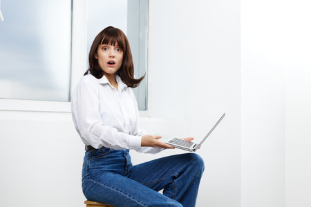 A surprised woman sits on a wooden stool by a bright window, using a laptop in casual attire. The...