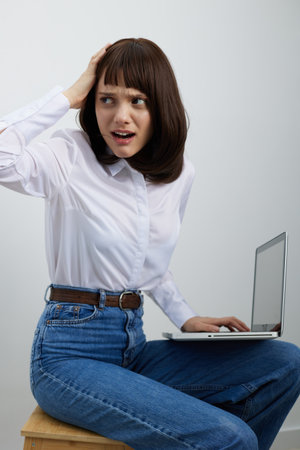 A young woman sits on a stool with a laptop on her lap, hand to her head, eyes wide in surprise as...