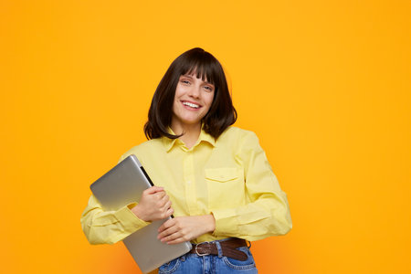 Cheerful young woman stands against a bright yellow backdrop, wearing a pale yellow shirt and jeans,...