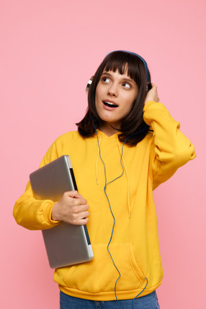 A young student wears a bright yellow hoodie and headphones while holding a laptop, set against a pink backdrop. The casual, cheerful scene conveys focus, energy, and a relaxed learning mood.の写真素材