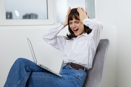 A surprised young woman sits in a bright home office, laptop on her lap, hands on her head, reacting...