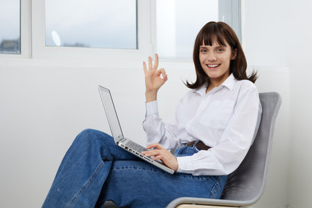 Bright, cheerful woman sits comfortably with a laptop in a modern home office, giving an OK gesture...