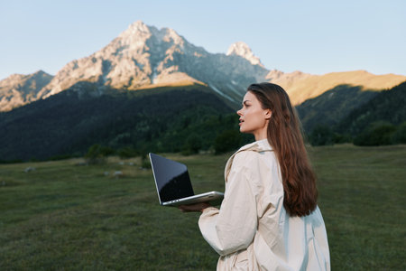A woman stands in a wide grassy valley with towering mountains in the background, holding a laptop and looking into the distance with a calm, focused expression.の写真素材