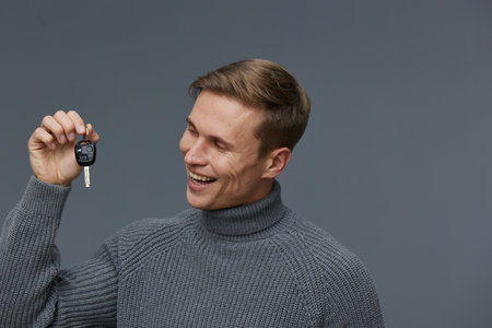 Smiling man holding car key with remote control in hand, wearing cozy gray turtleneck sweater,...