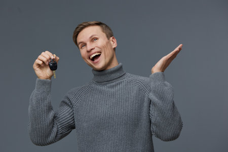 Cheerful young man with short brown hair wearing a cozy gray turtleneck sweater holding key in hand, smiling widely, looking excited, standing against plain gray background. Studio portrait, happy lifestyle concept.の写真素材