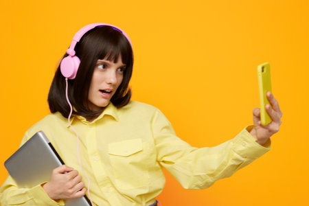 A cheerful young woman in a yellow shirt wears pink headphones and holds a tablet while taking a selfie against a bold orange background.の写真素材