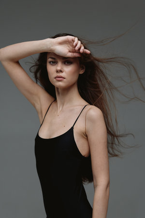 Young woman with long hair in black dress posing with hand on forehead against gray background