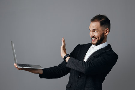 A professional man in a business suit holds a laptop, smiling and gesturing in triumph against a solid colored backdrop, conveying confidence, focus, and dynamic energy.の写真素材