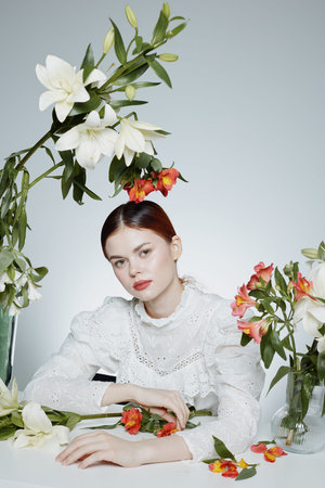 Beautiful young woman with red lips wearing a white blouse surrounded by fresh flowers on a light background, representing femininity and elegance in a serene settingの写真素材