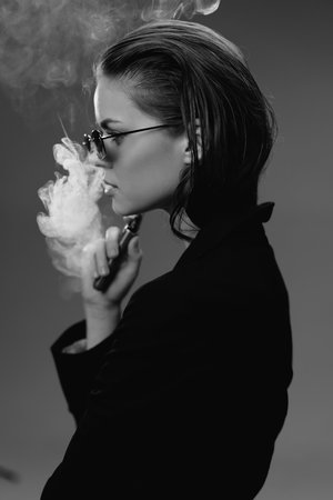 Black and white portrait of a woman vaping with glasses in a dark outfit exhaling smoke indoors.の写真素材