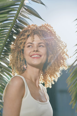 smiling woman with curly hair outdoors surrounded by green palm leaves, wearing white tank top, bright natural sunlight enhancing a cheerful summer vibeの写真素材
