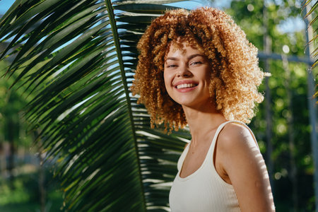 smiling woman with curly hair wearing white tank top stands outdoors near tropical palm leaves in warm sunlight, natural beauty and happiness captured.の写真素材