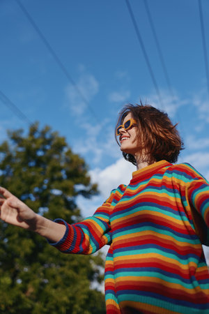 Woman in colorful sweater wearing sunglasses and smile outdoors under blue sky, portrait of a happy young adult enjoying a sunny day among trees, casual fashion and freedom.の写真素材