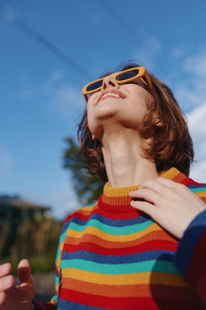 Woman smiling in striped sweater and orange sunglasses, short hair and looking up at sunny sky outdoors. Young adult enjoying travel lifestyle, joyful relaxed pose in warm light.の写真素材