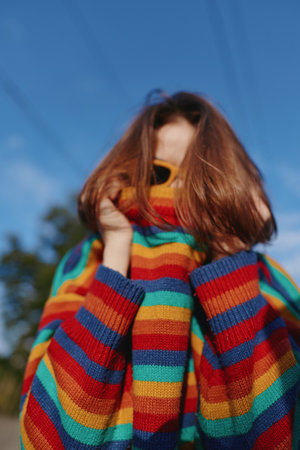 Young woman in her early 20s with red hair and sunglasses pulling a colorful striped sweater up over...