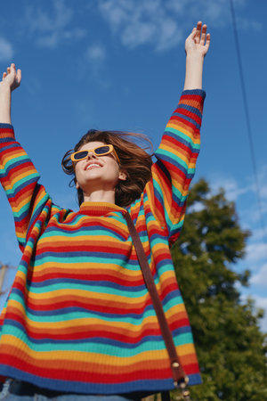 Woman in her 20s wearing a rainbow sweater and sunglasses, smiling with arms raised outdoors under blue sky, youthful lifestyle portrait, casual colorful fashion for travel and leisure.の写真素材