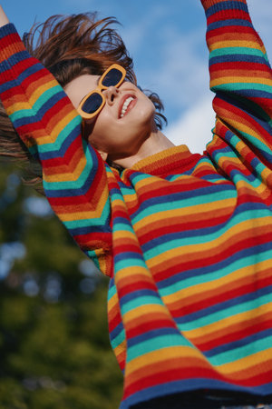 young woman in striped sweater and sunglasses smiling with arms raised outdoors under blue sky,...