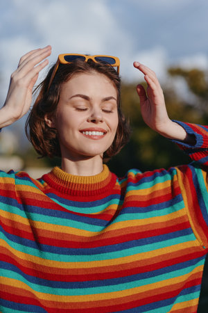 Woman portrait smiling with eyes closed wearing sunglasses on head and colorful sweater outdoors in sunlight. Happy young woman enjoying warm day and relaxed moment.の写真素材