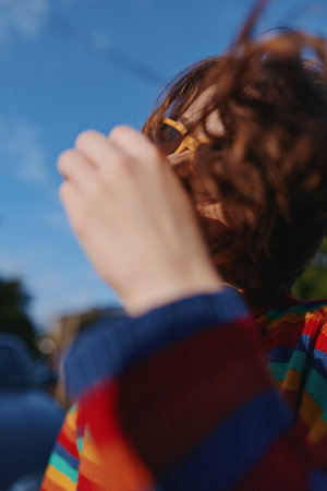 Young woman with curly hair in a colorful striped sweater wearing yellow glasses, candid smile and hand to face, outdoor portrait in sunny urban setting for lifestyle fashion.の写真素材