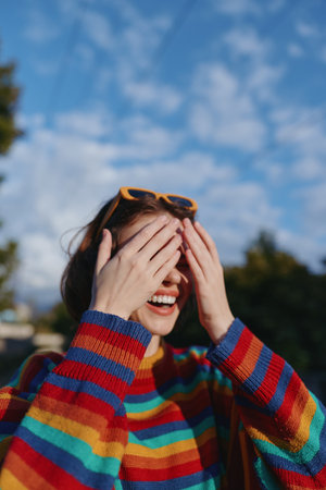 Woman smiling in a colorful striped sweater covering eyes with sunglasses on her head outdoor, laughing candidly in sunny blue sky setting, joyful mid-twenties lifestyle portrait.の写真素材