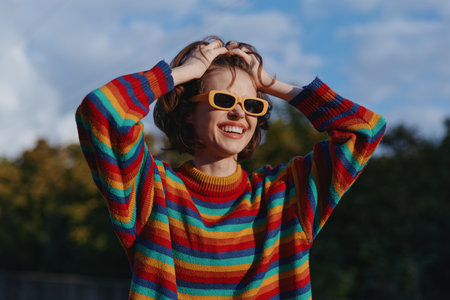 Woman in her 20s wearing a colorful striped sweater and yellow sunglasses, smiling with hands in hair outdoors in a park, casual outfit, joyful fashion lifestyle and warm autumn light.の写真素材