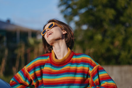 Young woman wearing a colorful striped sweater and yellow sunglasses, smiling and leaning back outdoors in a sunlit portrait, short brown hair and joyful expression for casual lifestyle fashion.の写真素材