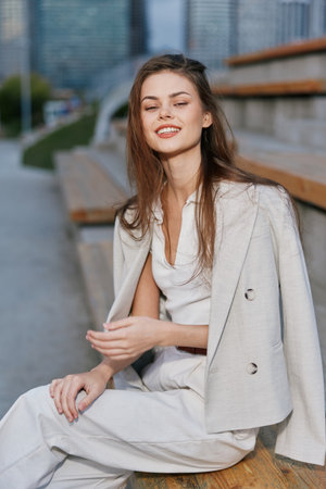 young woman in light business casual outfit sitting outdoors smiling serene confident urban background autumn fashion BUSINESSの写真素材