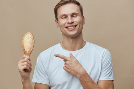 Smiling man holding hairbrush pointing to his head on beige background, casual clothing, cheerful expression, studio portrait, people lifestyle conceptの写真素材