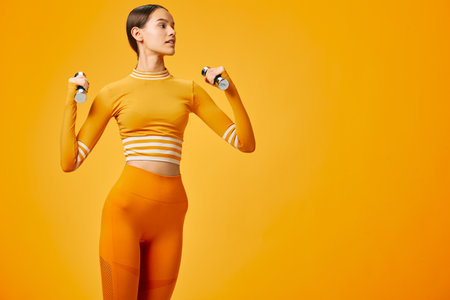 Woman fitness and sport theme: fit young woman in matching orange activewear holding light dumbbells, performing a shoulder exercise in a minimal studio with a vibrant orange background.の写真素材