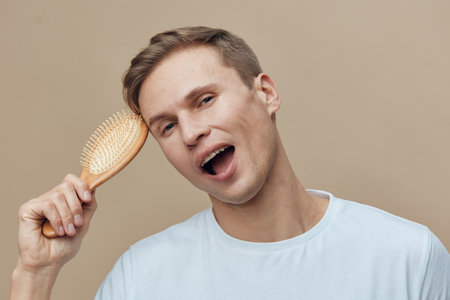 Smiling young man with short hair and fair skin holding a wooden hairbrush near his head, looking cheerful and relaxed in a casual light blue t-shirt. Studio portrait on a plain beige background, lifestyle concept.の写真素材