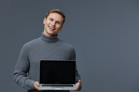 Smiling man holding open laptop computer in studio, casual attire, cheerful expression, modern technology concept, grey background, young adult male, leisure activity, technology use, digital lifestyle.の写真素材
