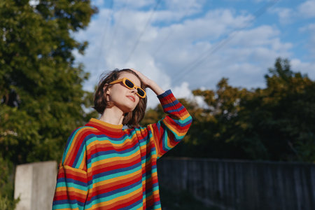 Woman in her 20s wearing a colorful striped sweater and sunglasses outdoors, smiling and looking up with hand on head, casual retro style in a sunny urban park lifestyle scene.の写真素材