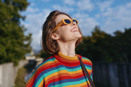 Young adult woman smiling, looking up while wearing yellow sunglasses and rainbow striped sweater outdoors in sunny urban park; colorful fashion, joyful travel lifestyle portrait.の写真素材