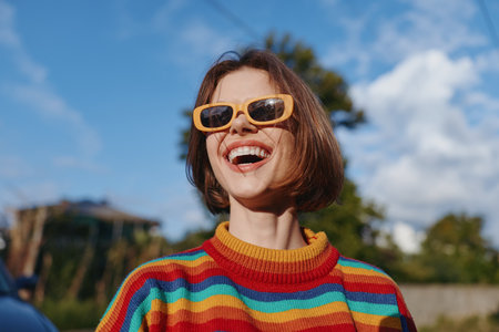 woman smiling striped sweater yellow sunglasses short hair outdoor sunny casual outfit happy lifestyle young adult laughing, wearing colorful knit, looking up in suburban park for travel and fashionの写真素材