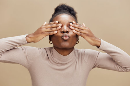 Happy African woman making playful faces while covering her eyes, wearing a beige turtleneck...